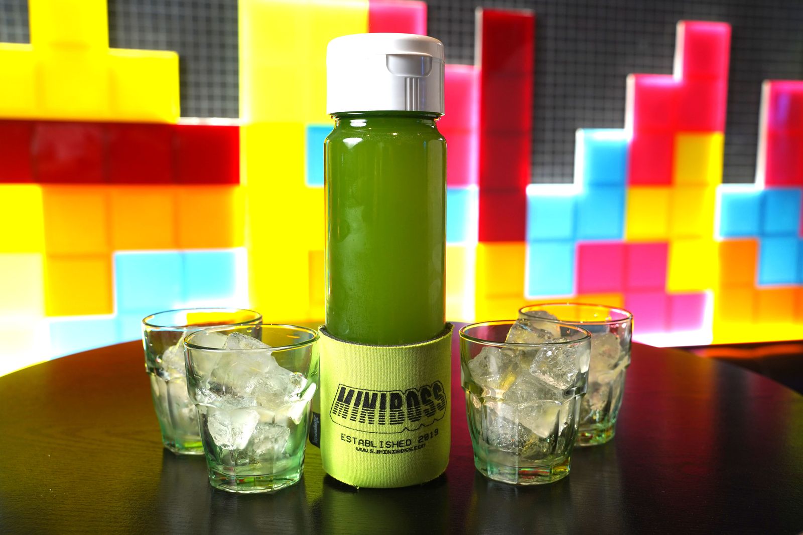 Bottle of green juice and small glasses on a table in front of a colorful block-pattern wall