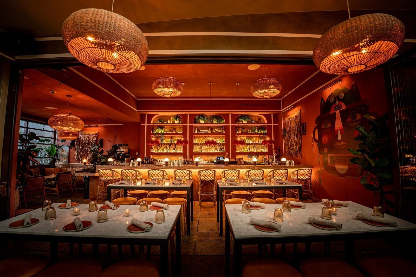 A dining room with white-topped tables and rattan pendant lights, facing a long bar lit in warm red tones.