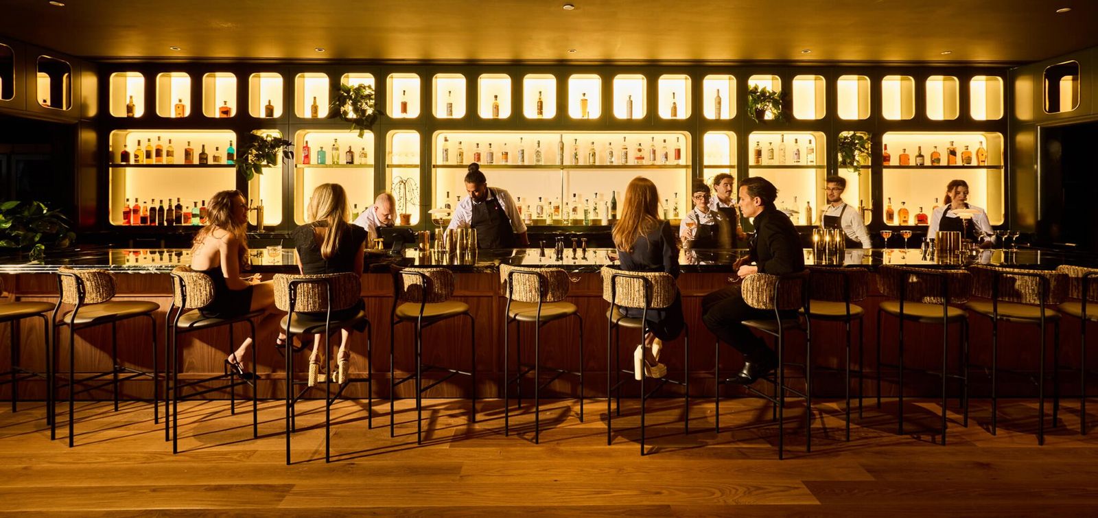 A long lit bar with a back wall of glowing bottle shelves and guests seated along the counter.