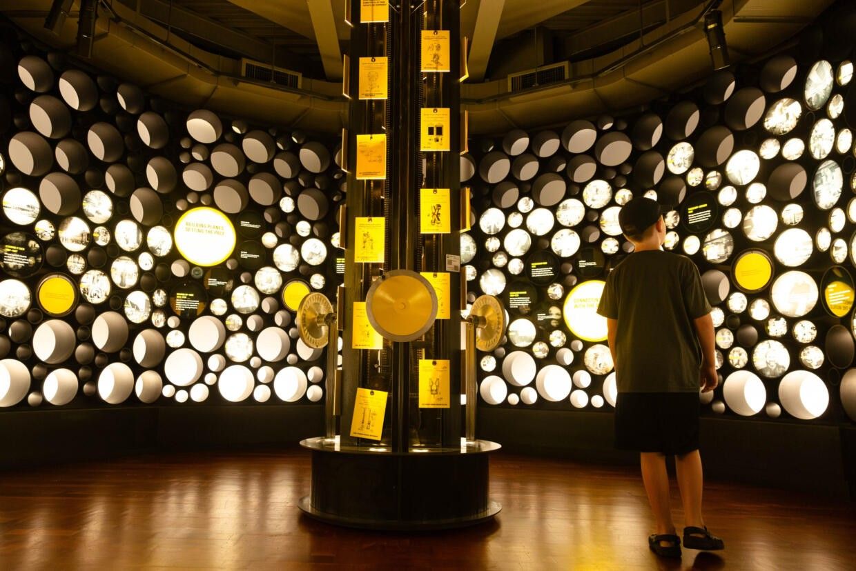 A dim gallery with a tall display column ringed by lit circular panels, with one visitor seen from behind.