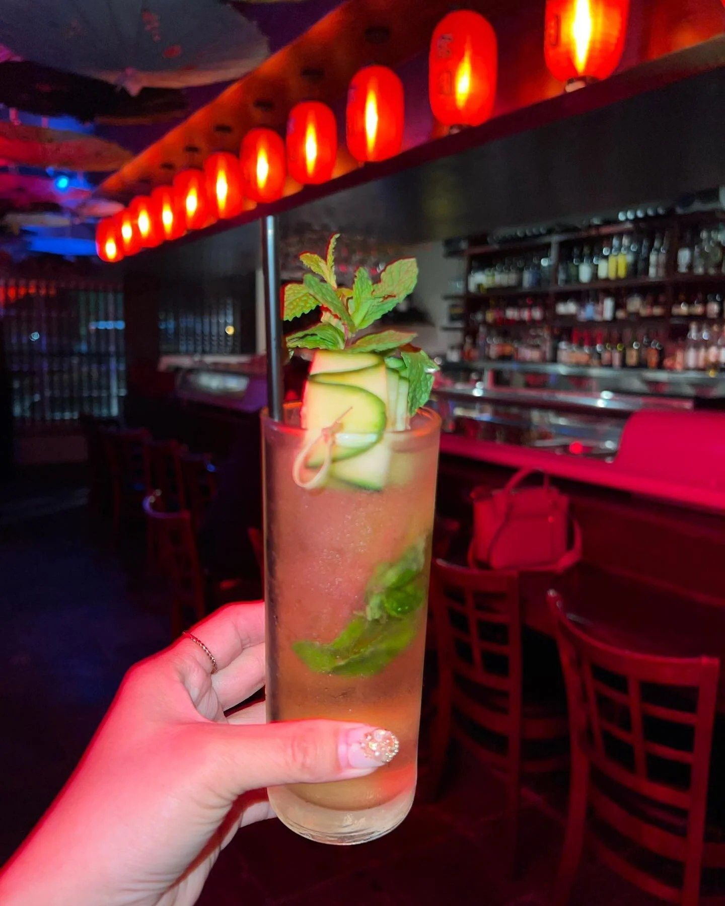 A hand holding a pink cucumber-and-mint cocktail in front of a bar lit with red lanterns.
