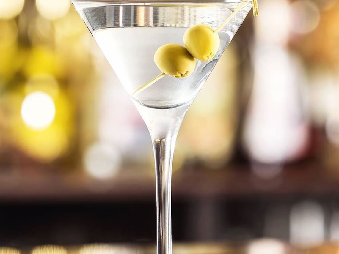 A martini in a stemmed glass with green olives on a pick, with warm lights blurred behind.
