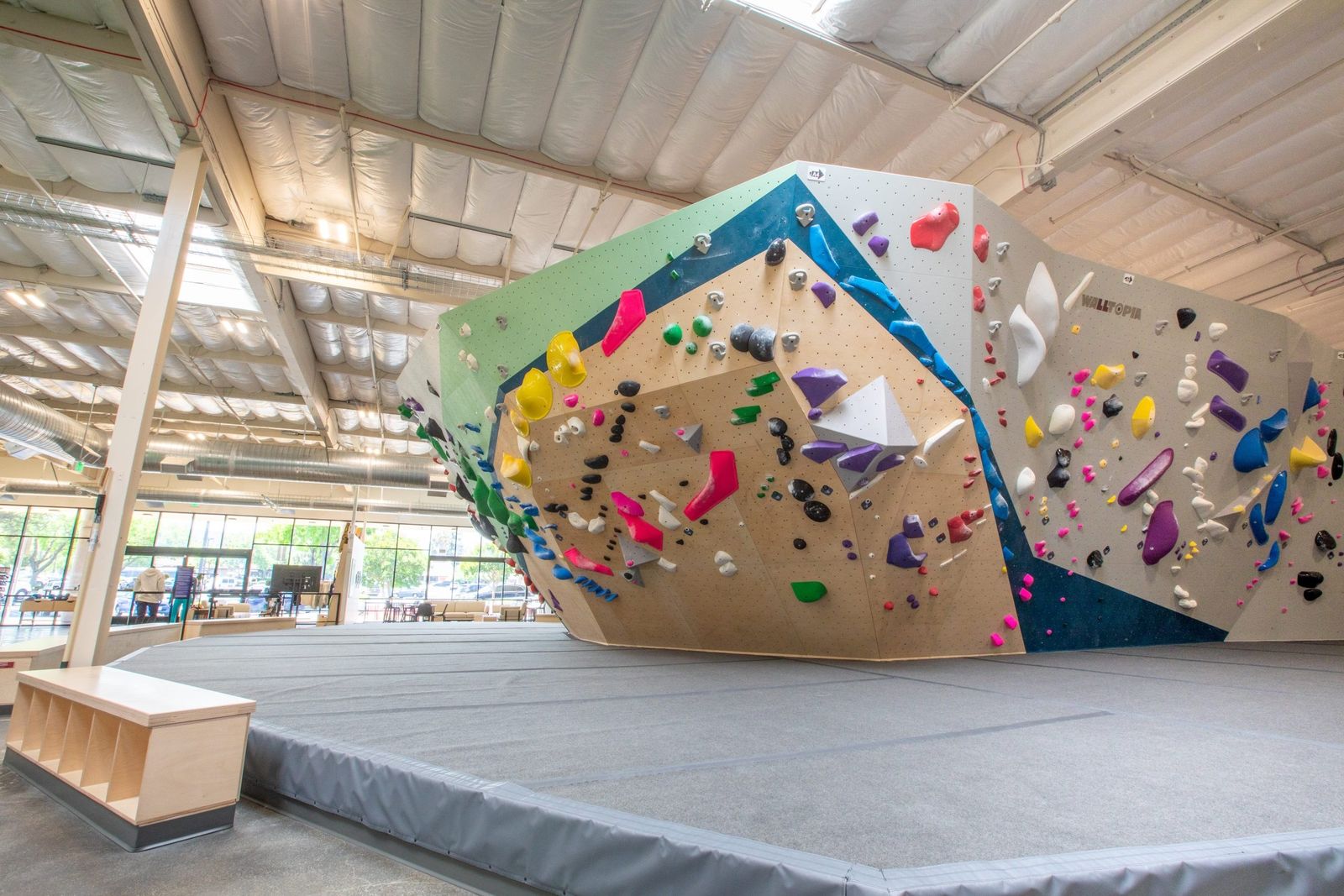 A bouldering wall with colorful holds and angled panels inside a high-ceilinged gym.