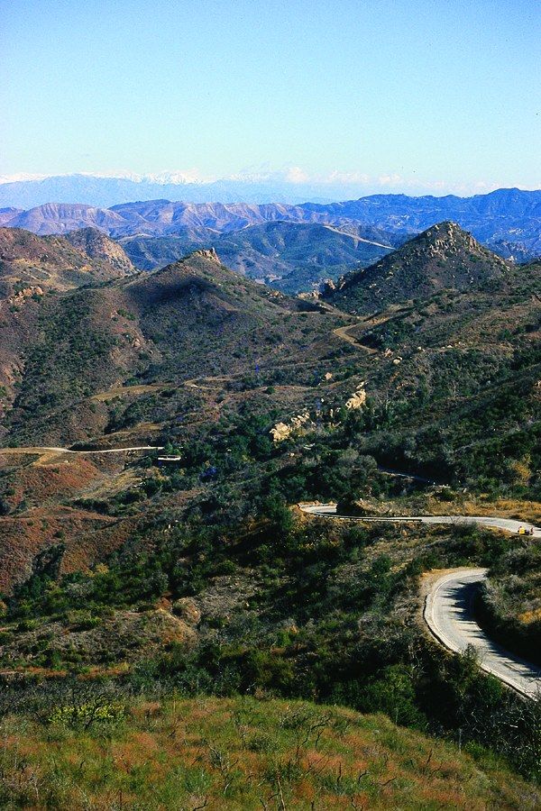 A winding mountain road through green hills and ridgelines under a clear sky.