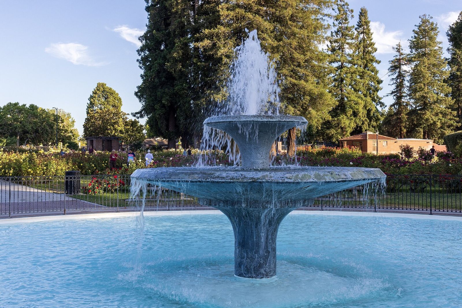 Stone fountain spraying water in a round pool with rose beds and tall trees behind it