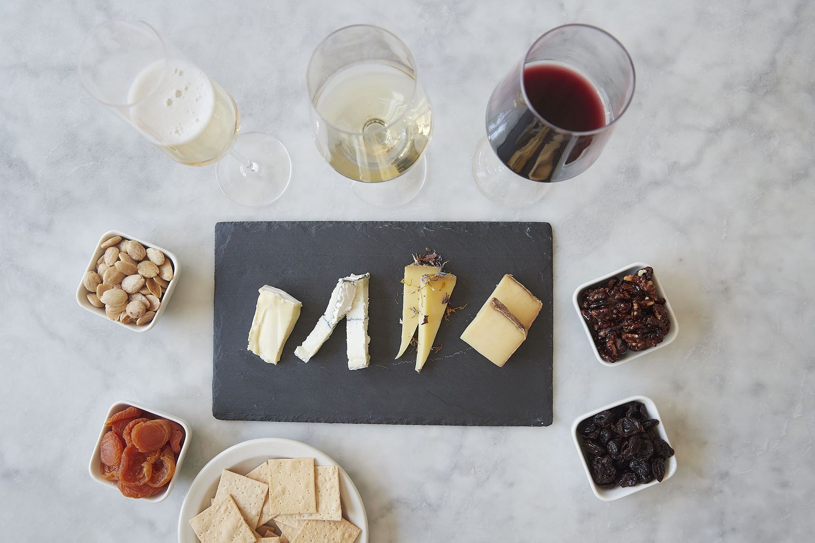 Overhead view of a slate cheese board with wedges, crackers, nuts and dried fruit beside glasses of wine