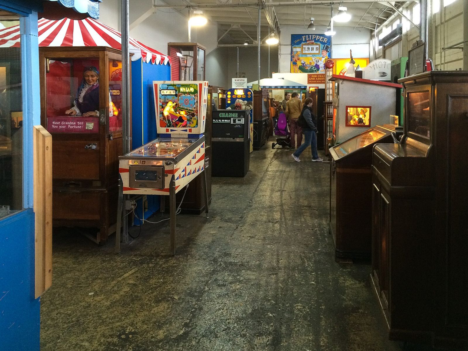 A dim arcade corridor lined with antique coin-operated machines and pinball games under a striped canopy.