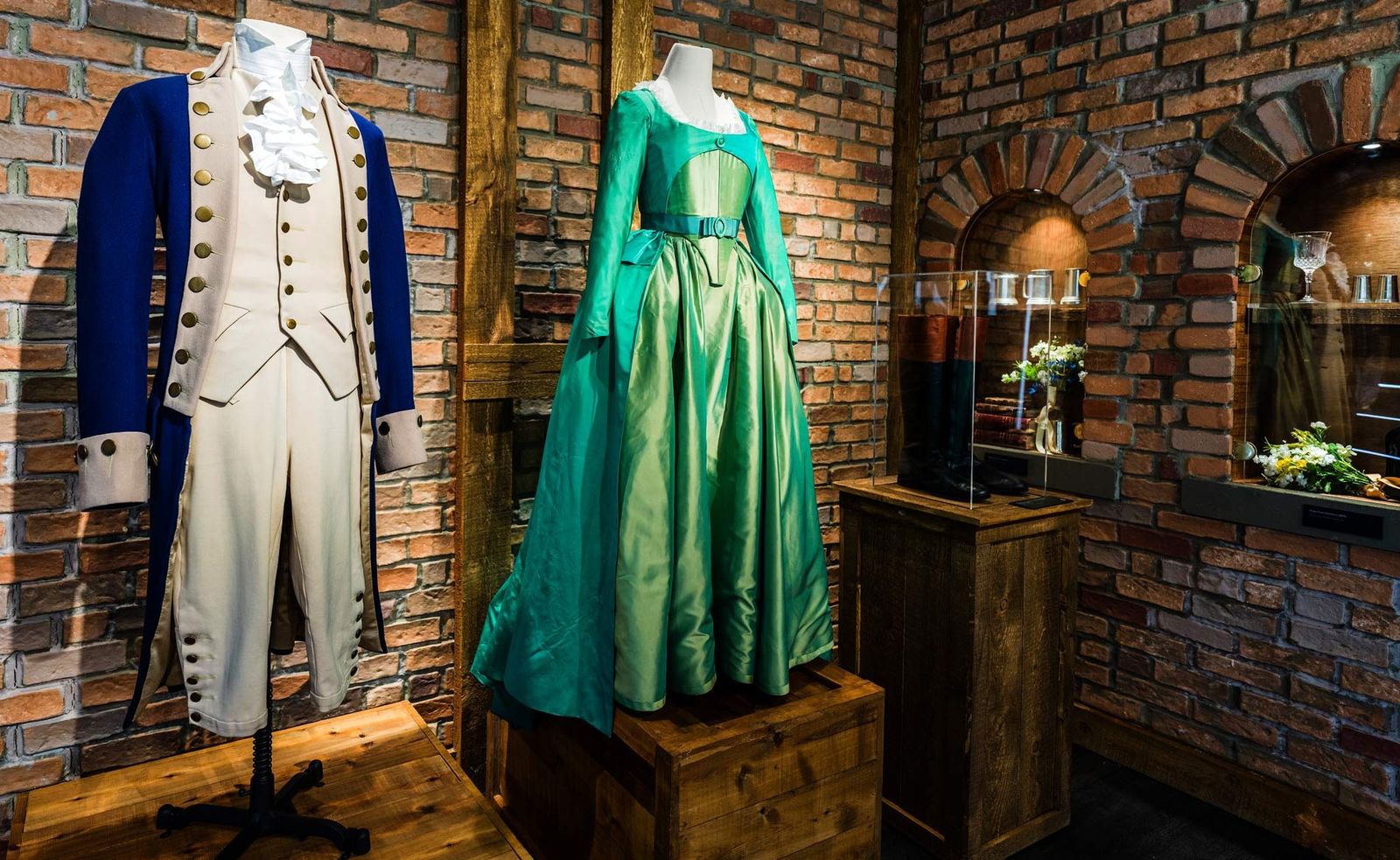 Two period costumes on mannequins, a blue coat with white breeches and a green gown, in front of a brick wall