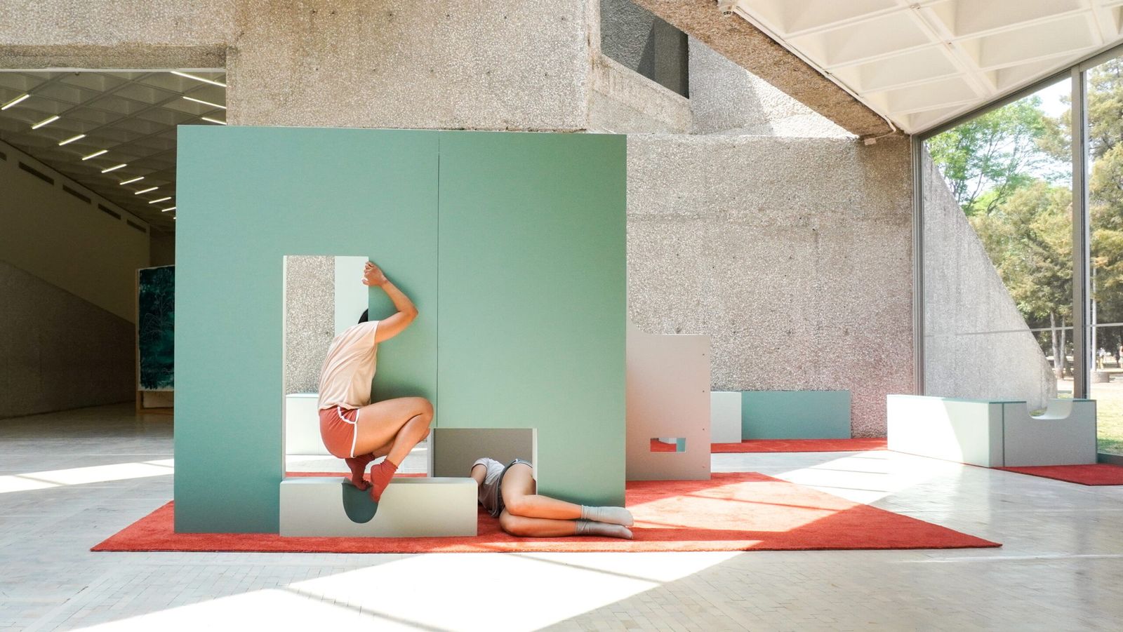 A bright gallery with a pale green sculptural installation and a person lying across it, beside a concrete wall and large windows.
