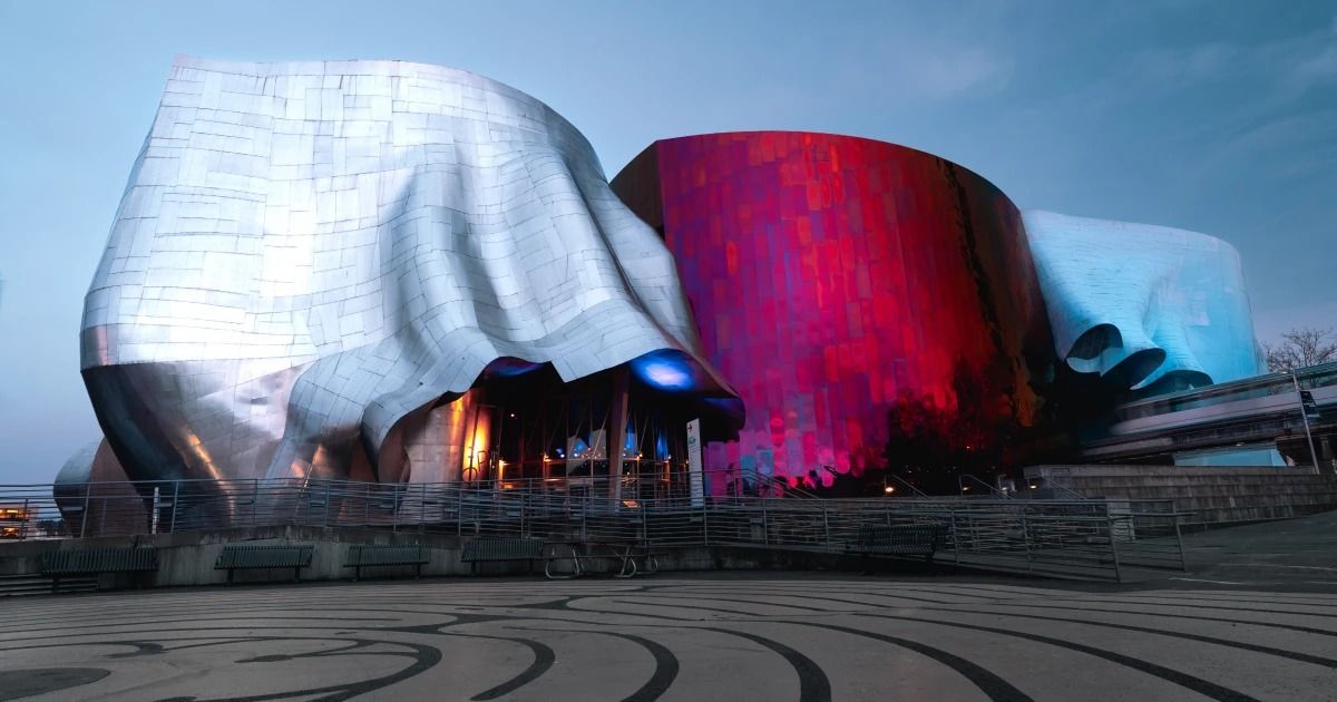 The museum's curving metal building in silver, red and blue panels at dusk, with a patterned plaza in the foreground.