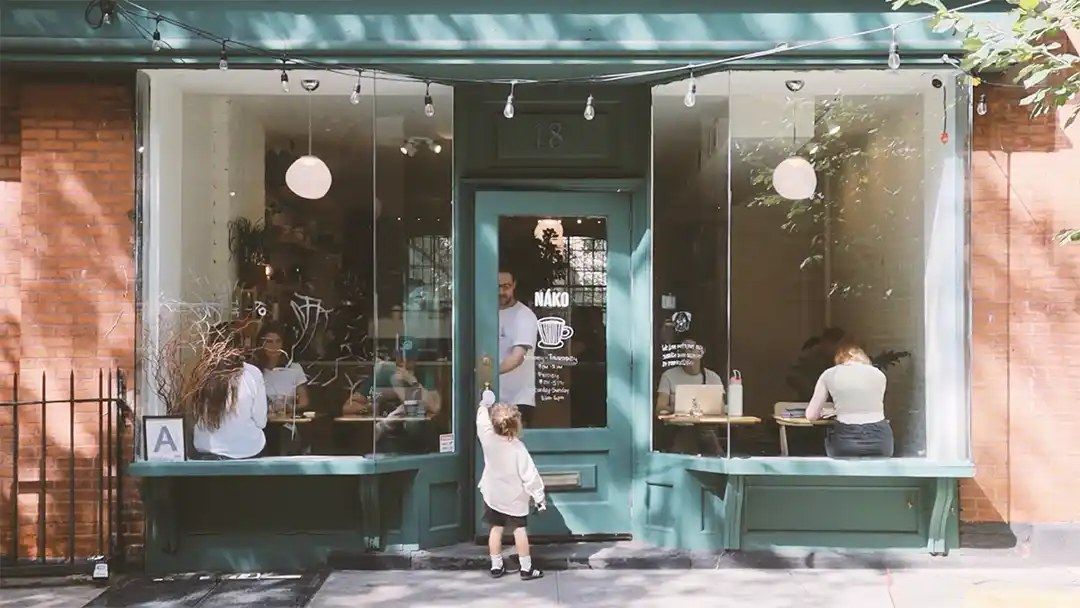 A teal-trimmed corner cafe with large windows, people seated at the window bench and a small child at the door.