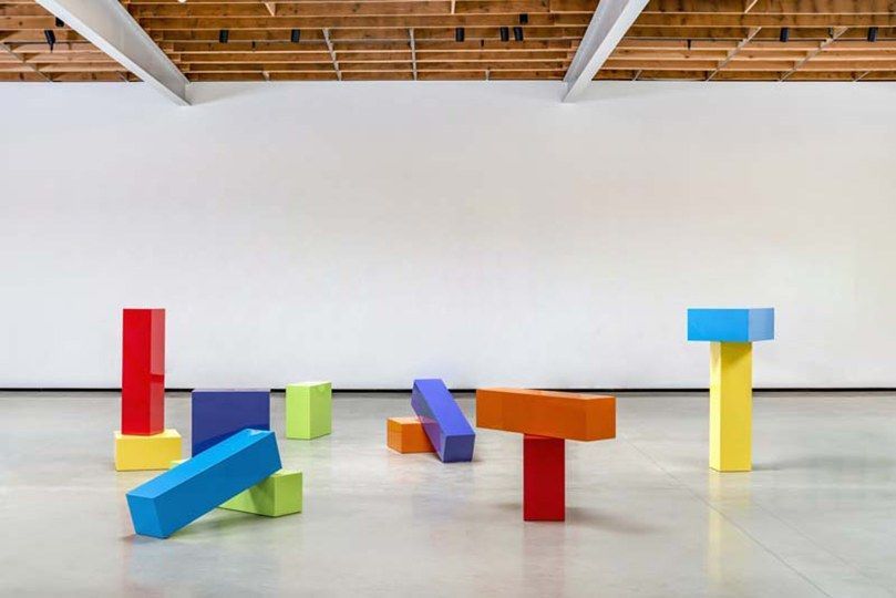 A white gallery with colorful geometric sculptures in red, blue and yellow on the floor.