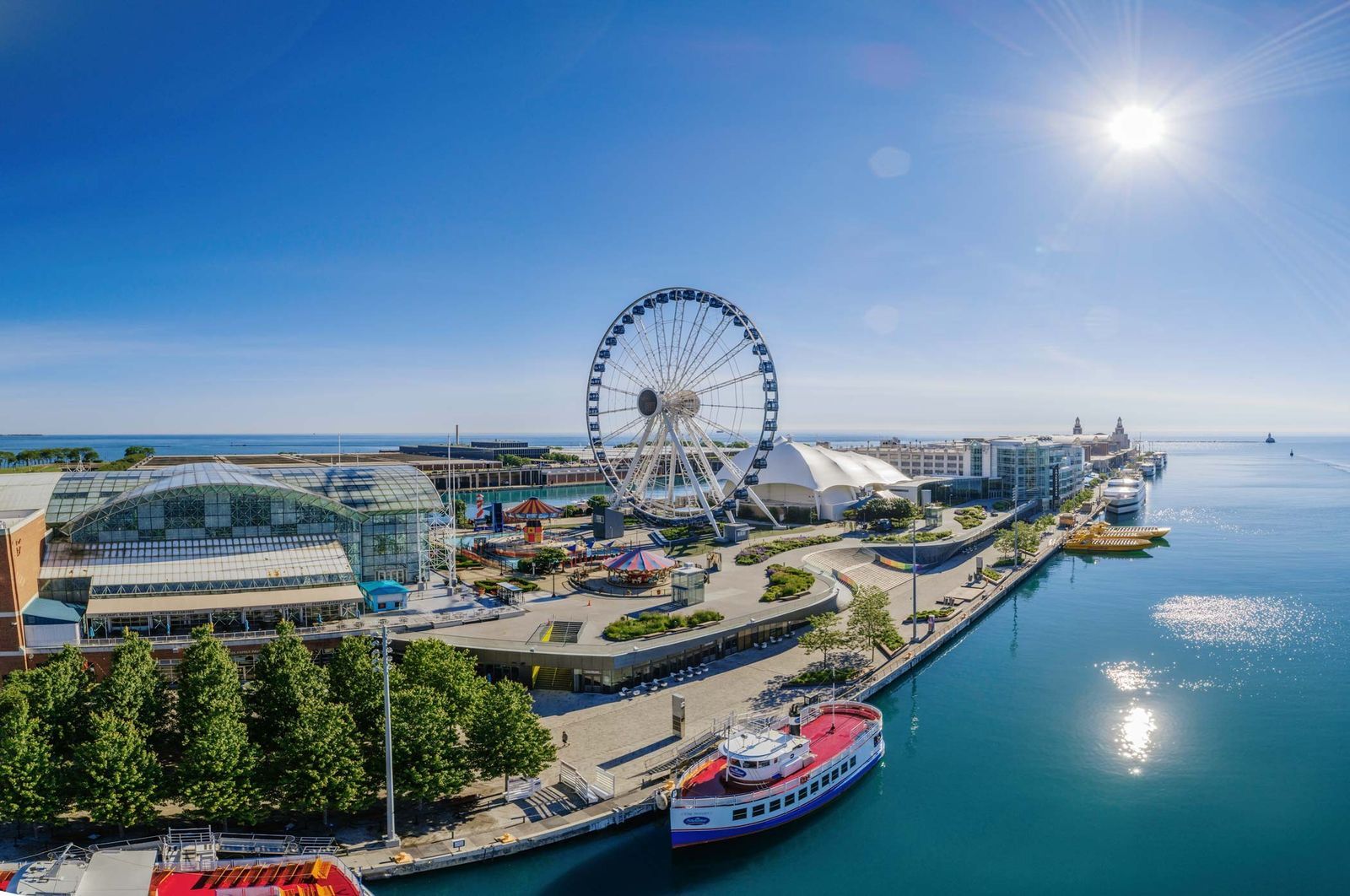 An aerial view of the pier with a Ferris wheel, buildings and boats on the blue lake under a bright sun.