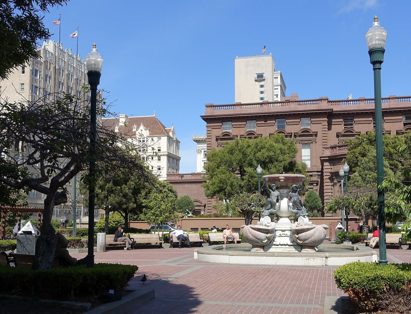A brick plaza with a tiered stone fountain, tall lamp posts and trees, with a large brown brick building behind.