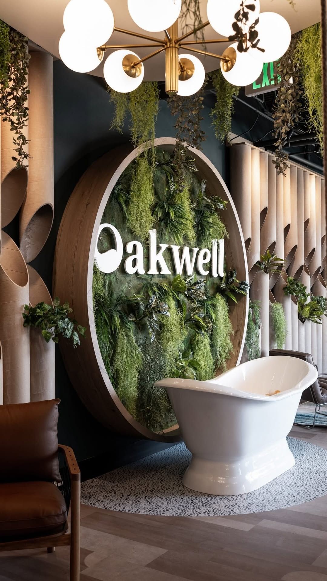 A round Oakwell sign over a white freestanding tub, with a wall of hanging greenery and a chandelier above.