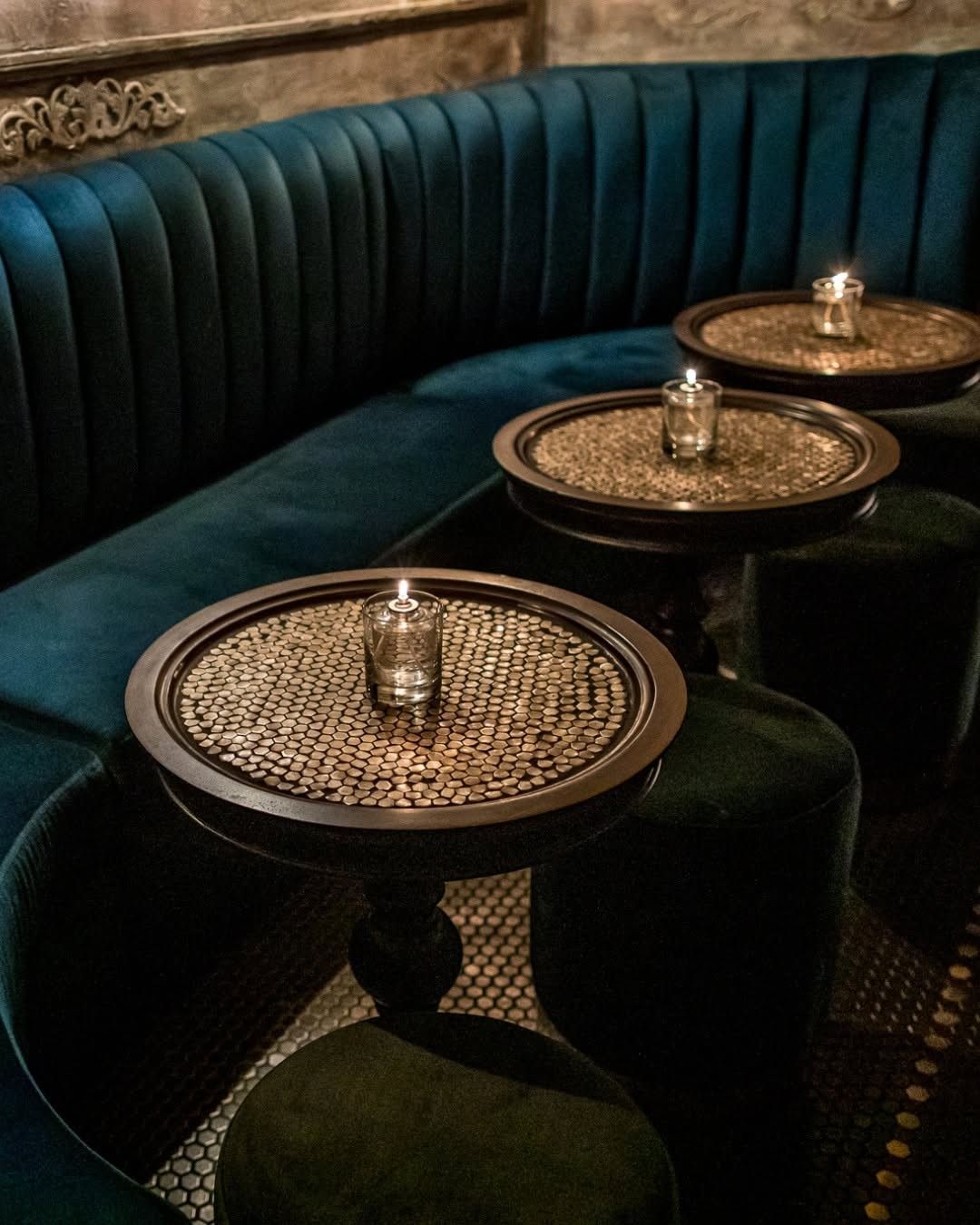 A curved teal velvet booth with small round brass tables and candles.