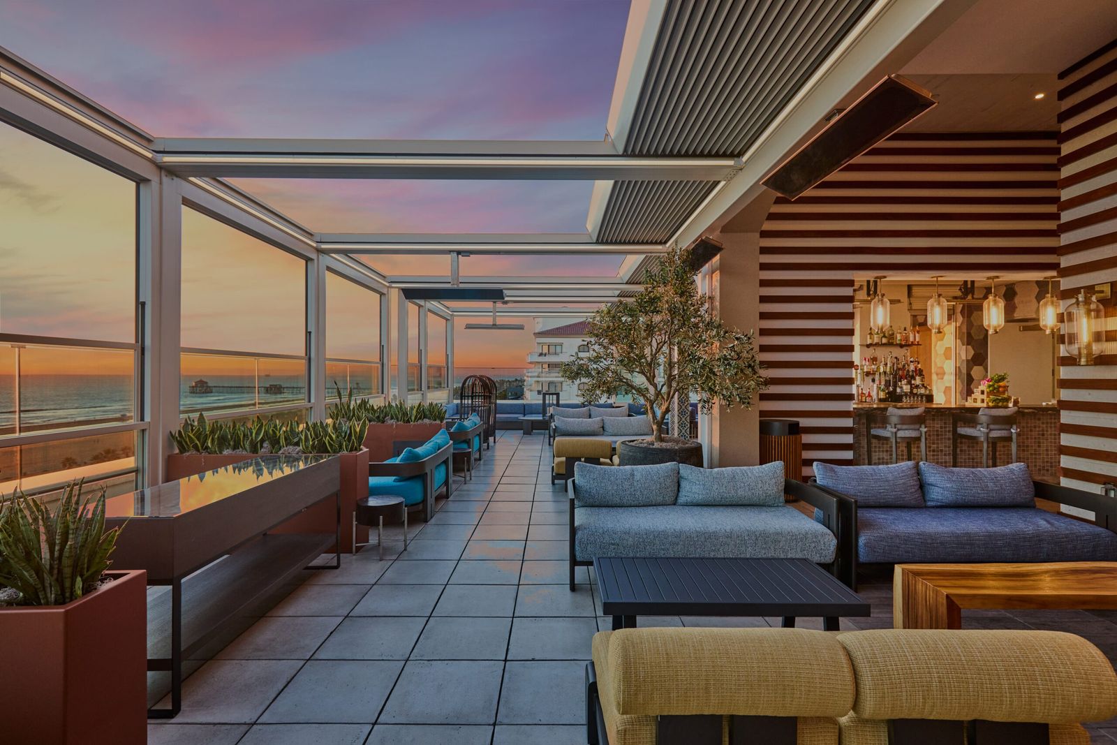 A rooftop lounge with sofas and loungers on a tiled deck at sunset, with glass railings.