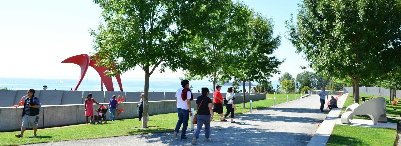 A paved waterfront path through the park, with a red steel sculpture on the left and a few people walking.