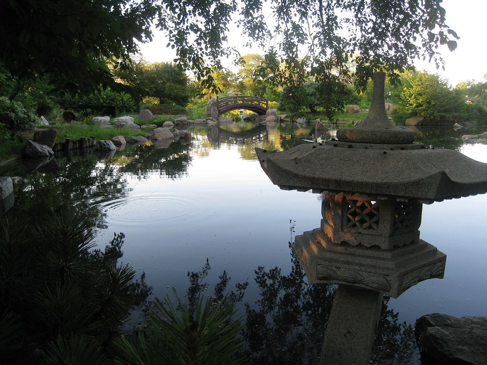 A stone lantern at the edge of a still pond with an arched bridge and trees behind.