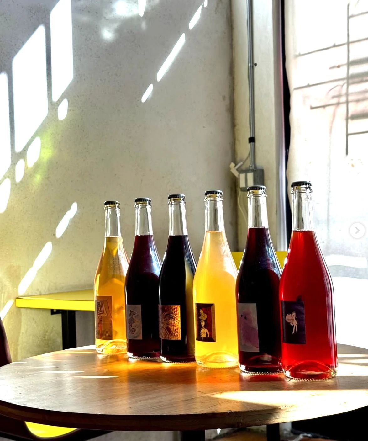 Six wine bottles in different colors lined up on a round wooden table in front of a concrete wall.