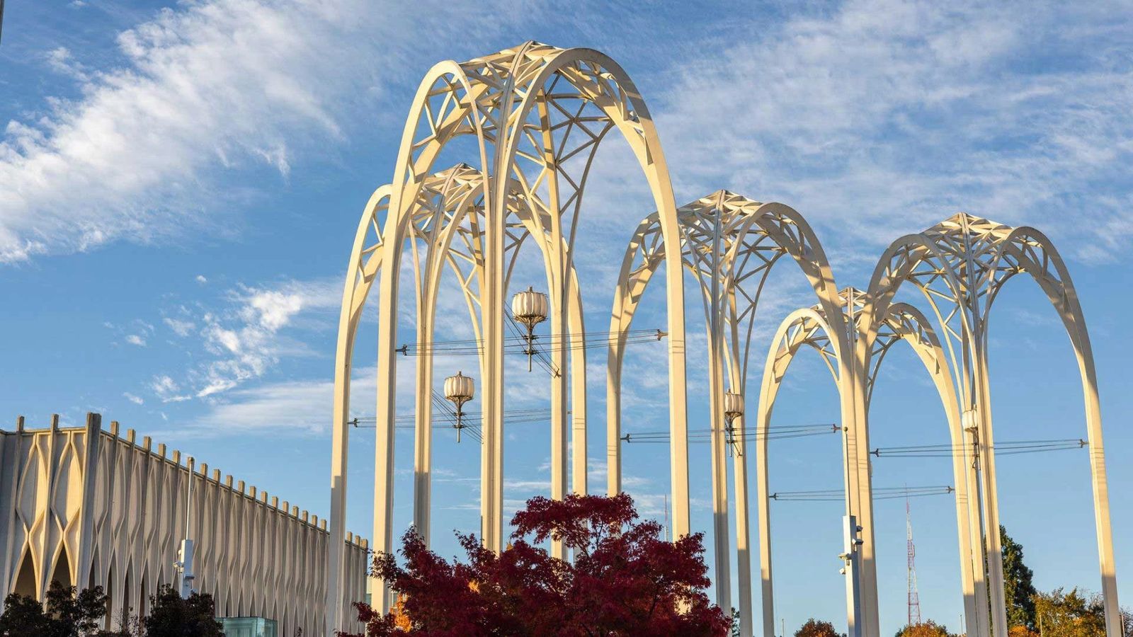 White arched towers rising against a blue sky with wispy clouds, beside a patterned white building, with a red-leafed tree below.