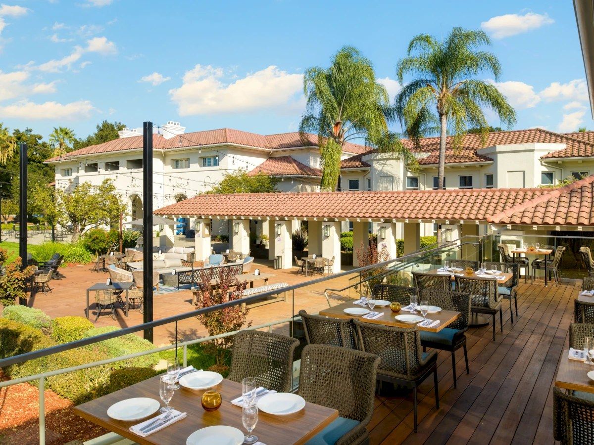 An outdoor restaurant patio with set wooden tables, a covered terrace and palm trees beyond