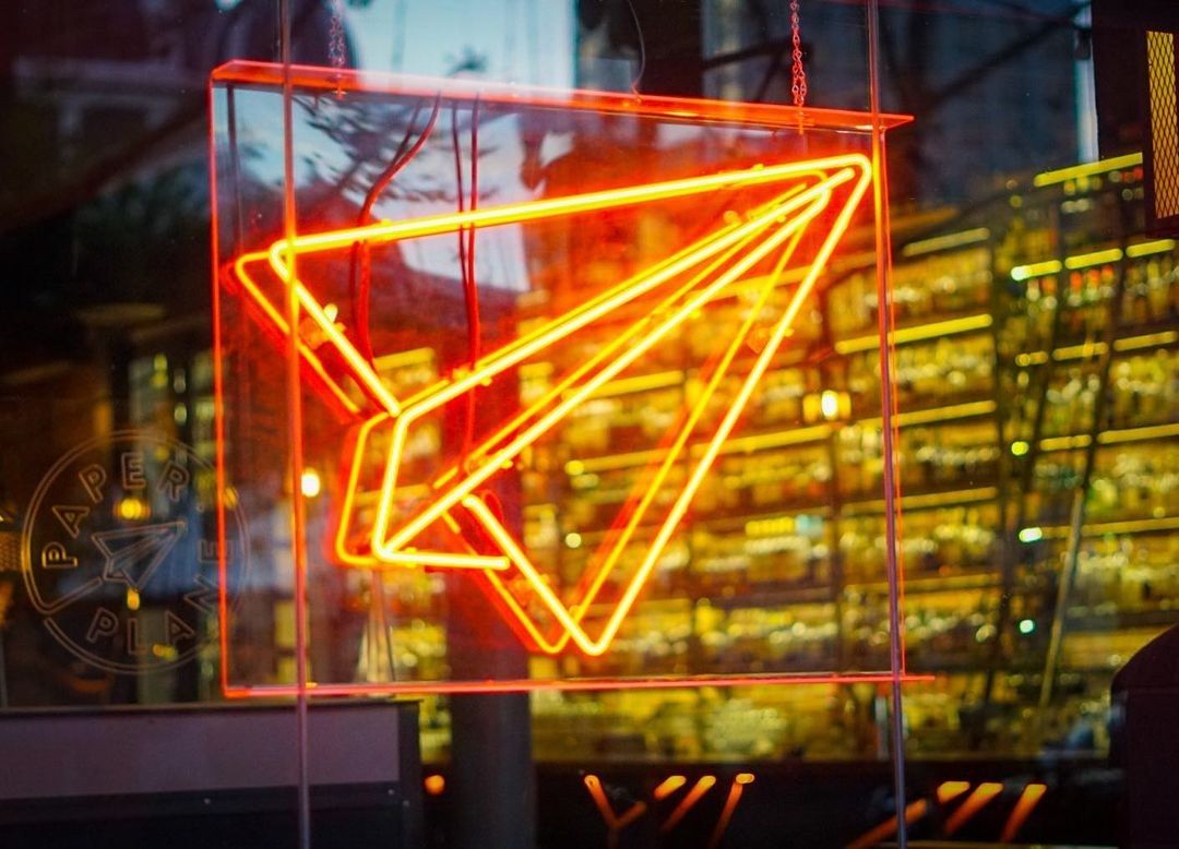 Glowing orange neon paper plane sign in front of shelves of bottles