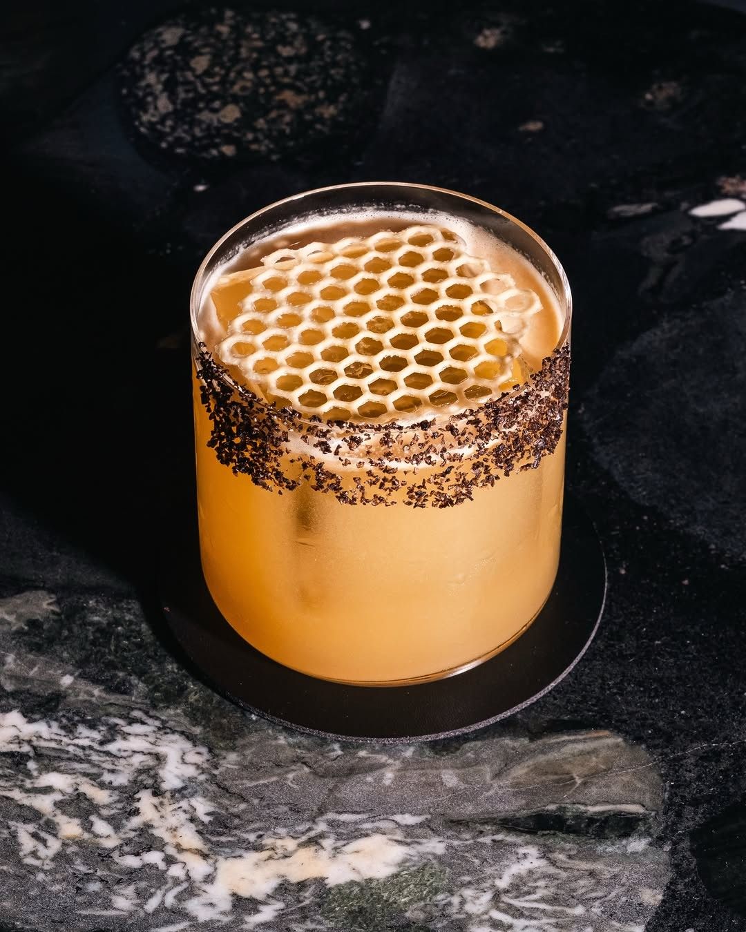 A cocktail in a short glass rimmed with spice and topped with a honeycomb, on a dark stone coaster.