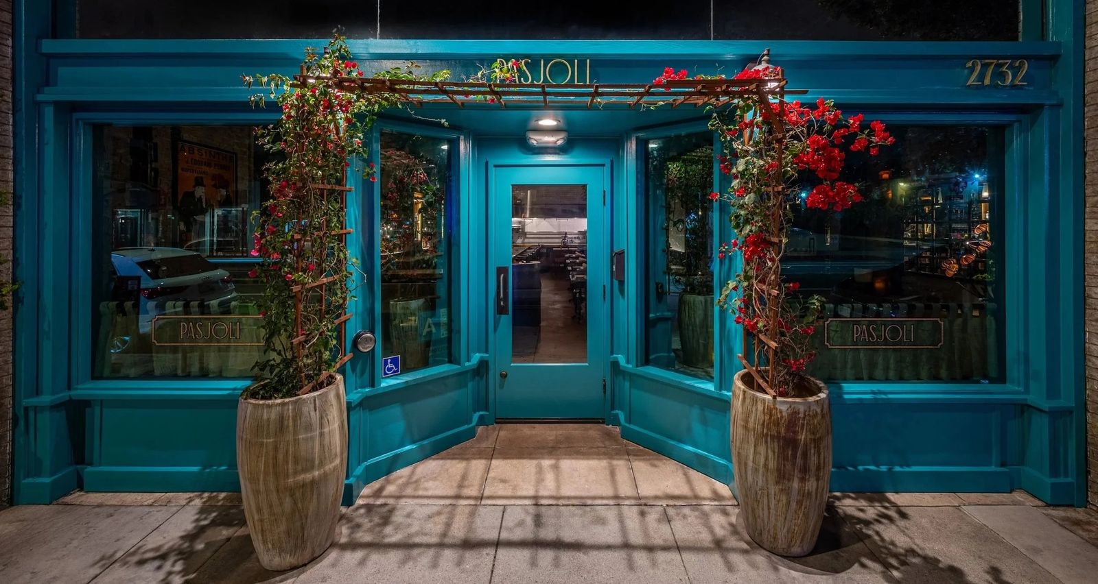Teal storefront of Pasjoli with flowering plants in barrels around the entrance, lit at night