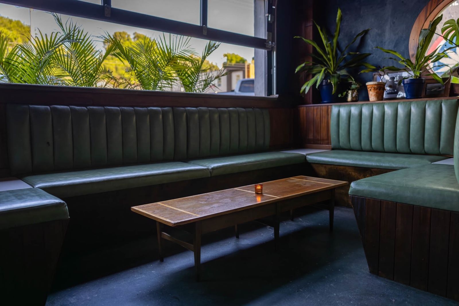 Green tufted booth seating and a low wooden table beside large windows with plants