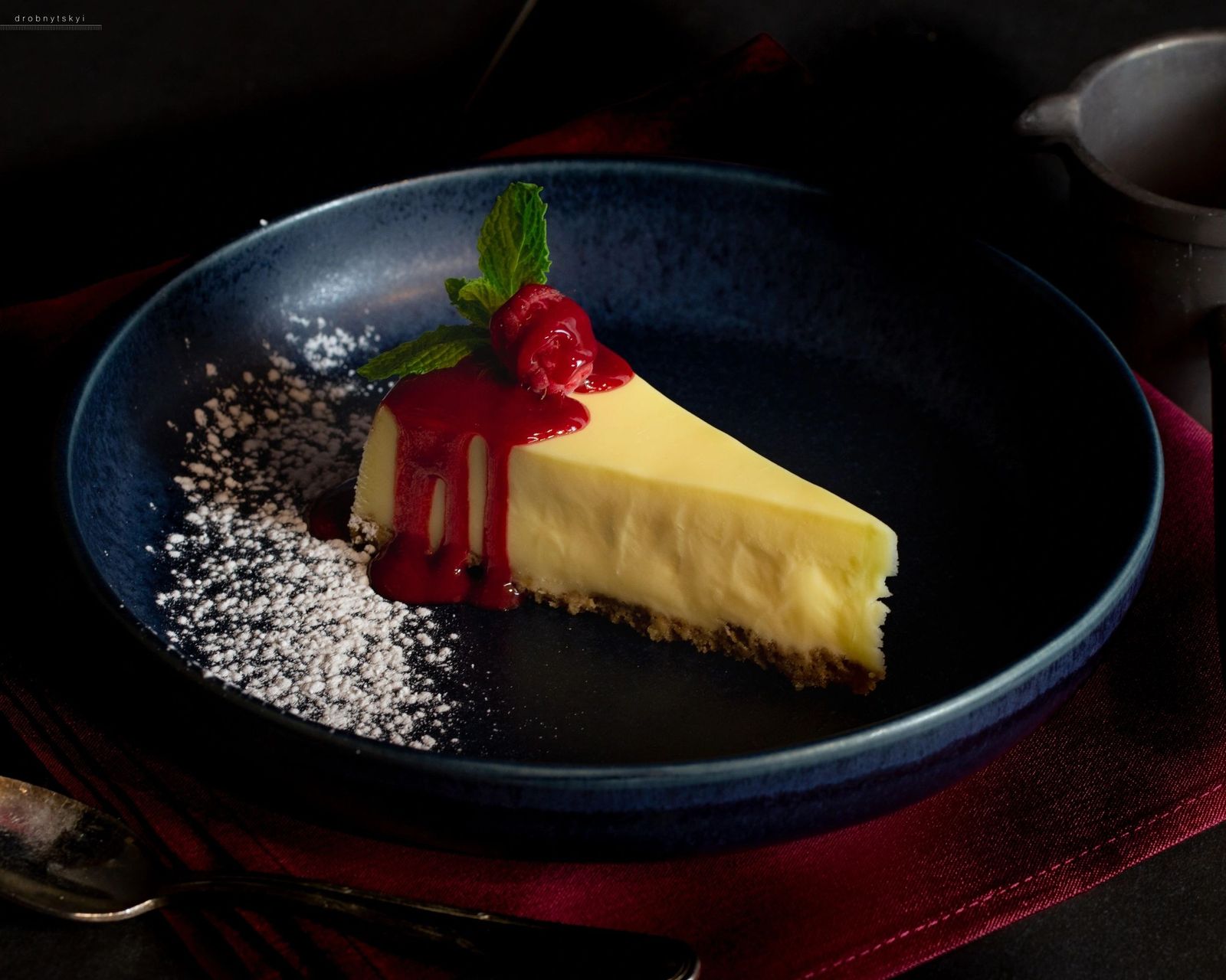A slice of lemon cheesecake with raspberry sauce and a mint leaf on a dark blue plate.