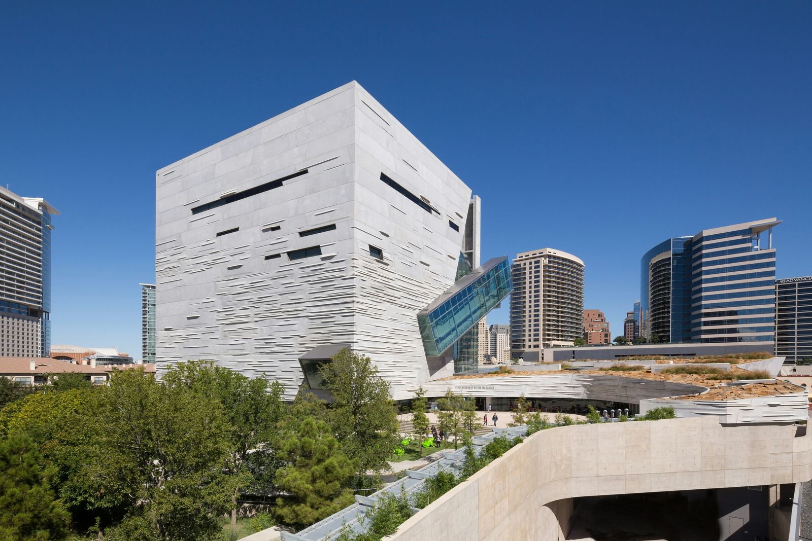 The museum's cube-shaped concrete building beside a green roof and city towers under a blue sky.