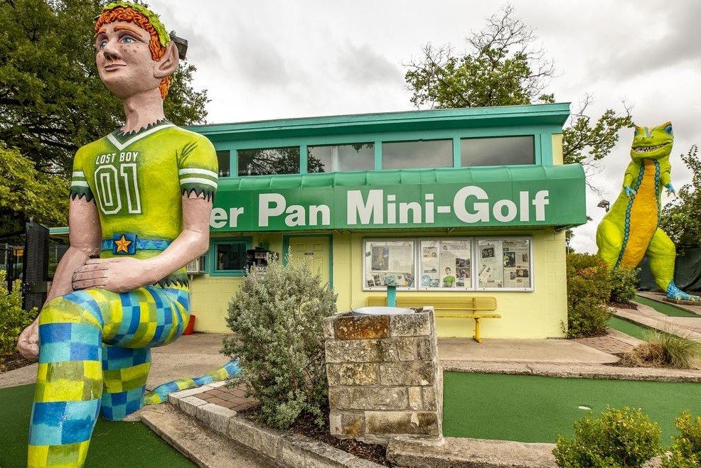 A statue of a boy in green and yellow in front of a small building with a Peter Pan Mini-Golf sign and a dinosaur figure.