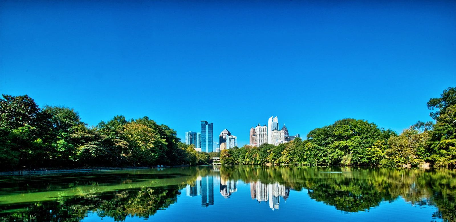A calm lake in a park reflecting trees, with the Atlanta skyline in the distance under a blue sky