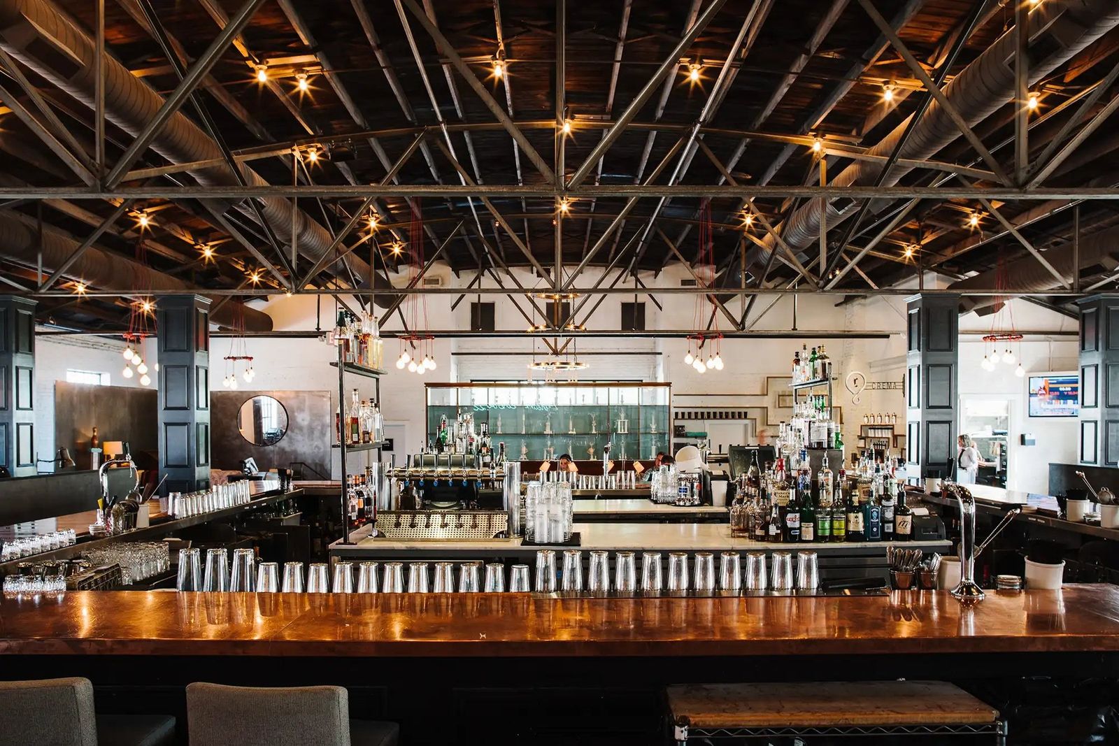 Long wooden bar with bottles and an industrial ceiling with exposed trusses