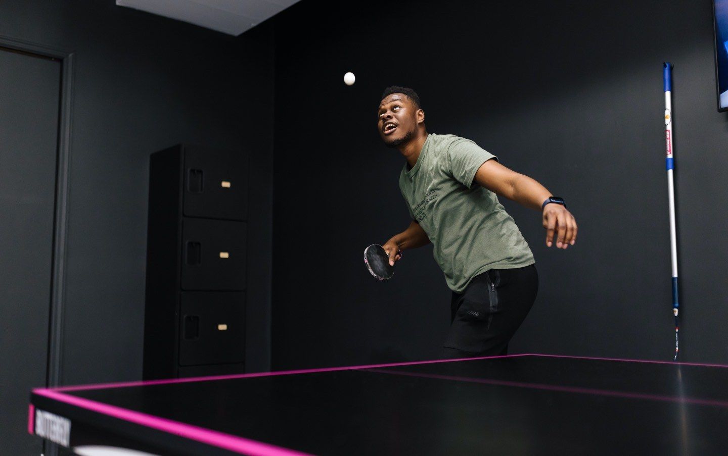 Player in a dark ping pong room mid-swing beside a black table with pink edge lines