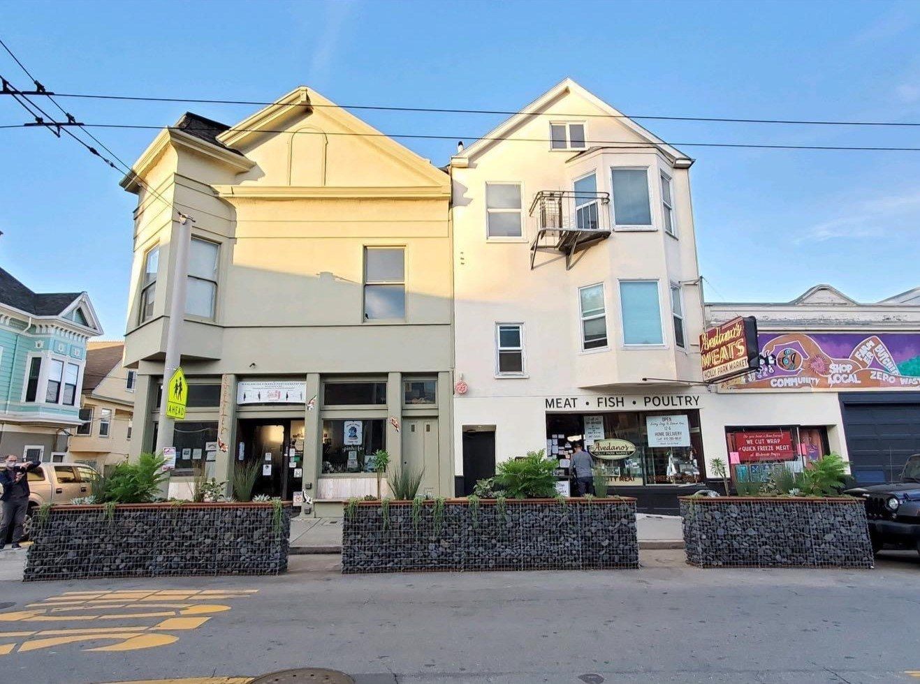 A cream-colored two-story corner building with a cafe at street level on a hill, with a few plants out front.
