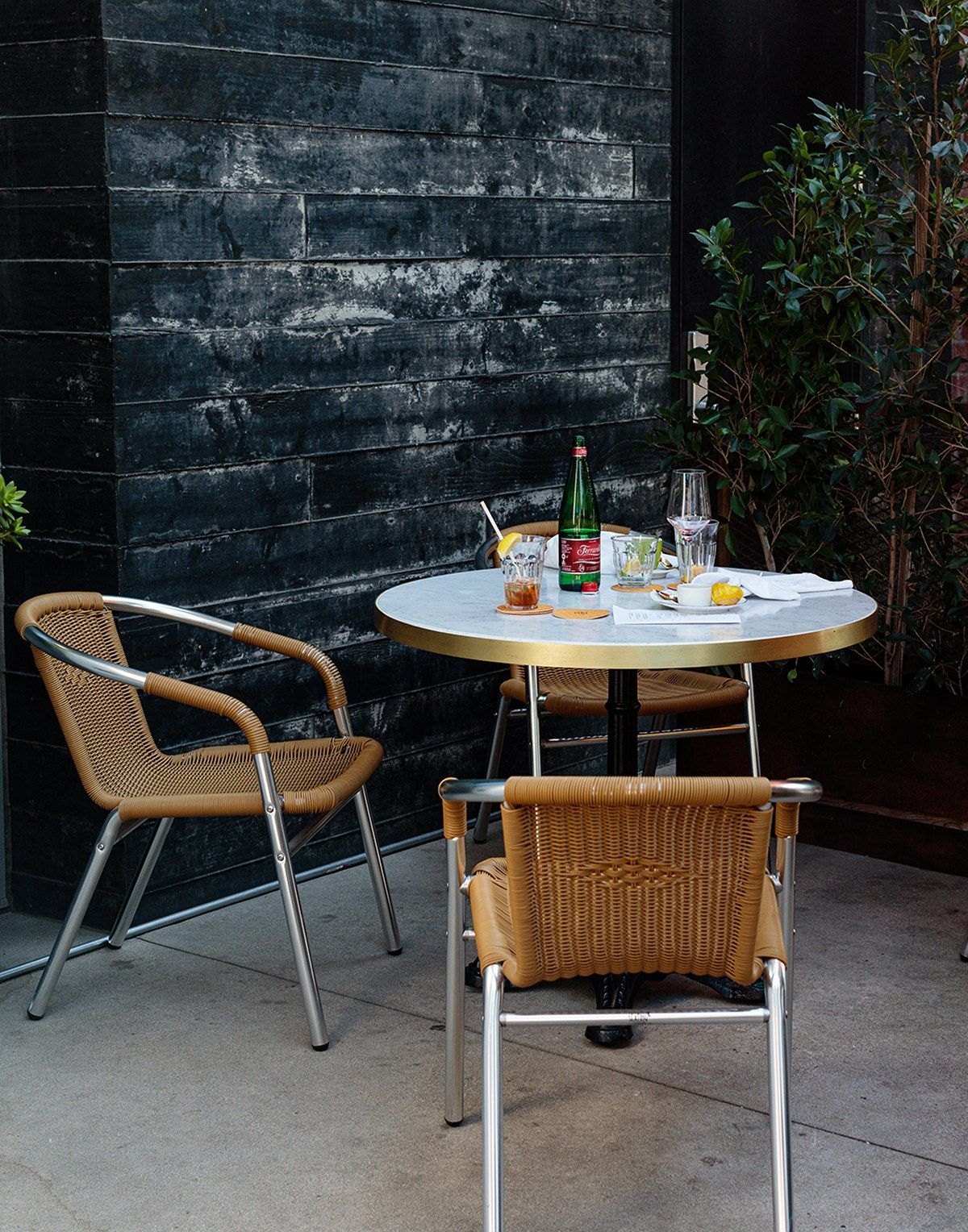 Round cafe table with drinks and two woven chairs against a dark stone wall in an outdoor courtyard