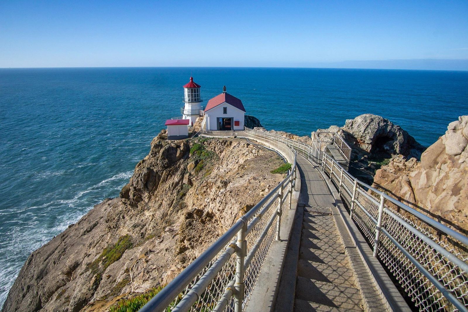 The Point Reyes lighthouse perched on a rocky headland above blue ocean, with a railed stairway leading down to it