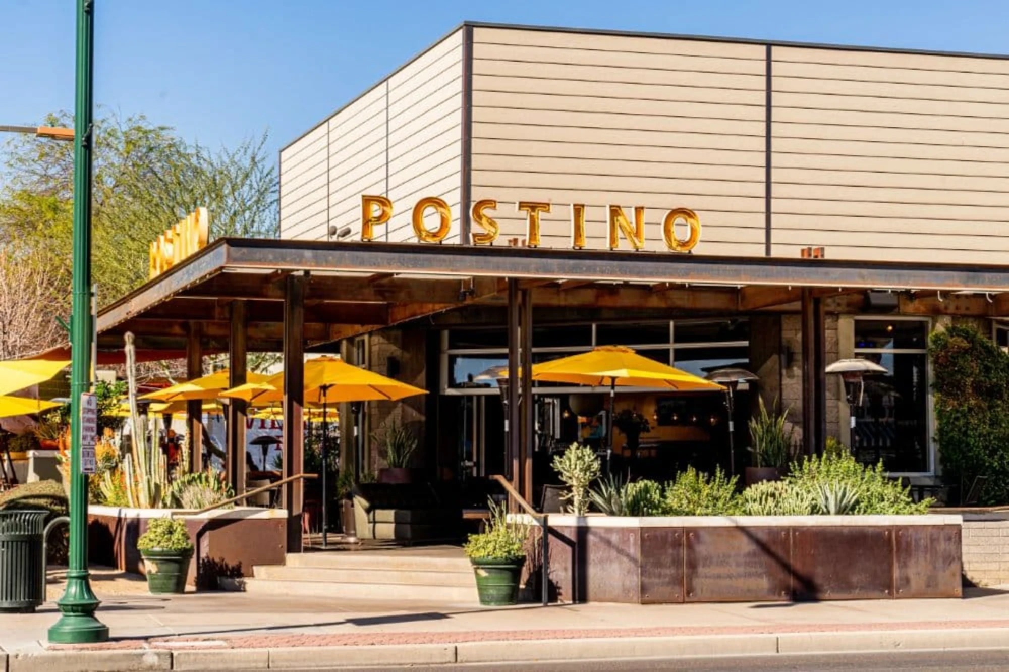 Postino WineCafe building with gold block lettering and a patio of yellow umbrellas and desert plants