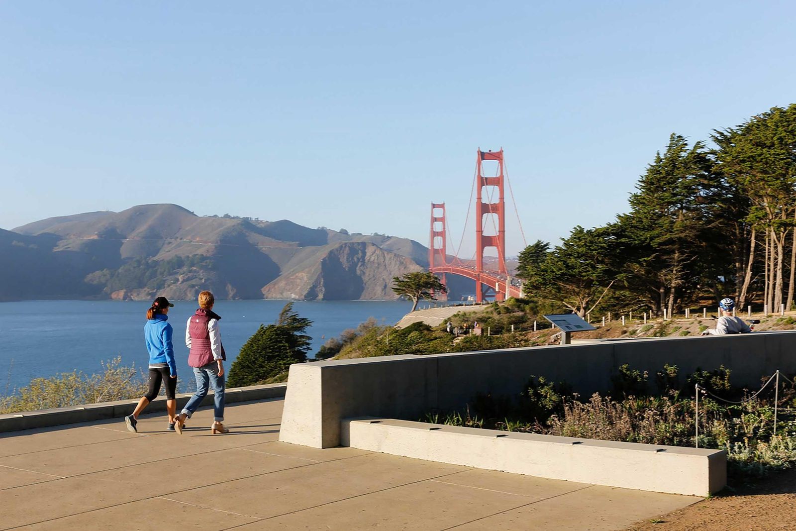 Two walkers on a paved overlook by a low concrete wall with the Golden Gate Bridge and headlands beyond.