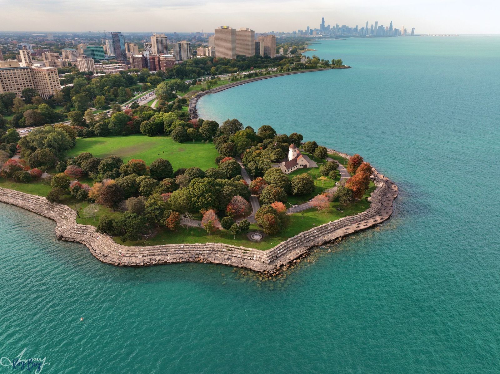 An aerial view of a tree-covered peninsula with a stone-edged shoreline and the city skyline beyond turquoise water.