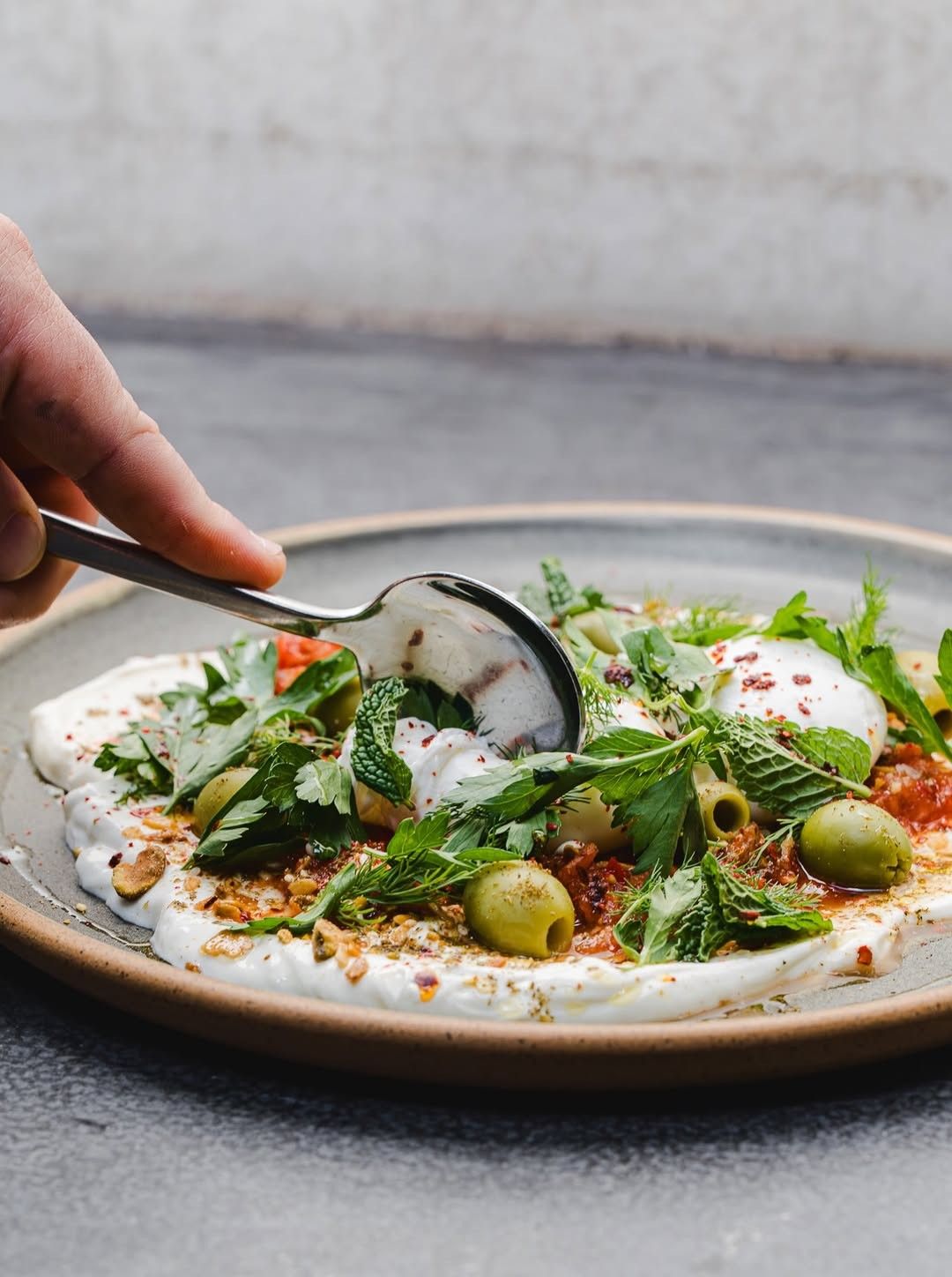 A plate of yogurt with greens, olives and olive oil, with a hand spooning from it.