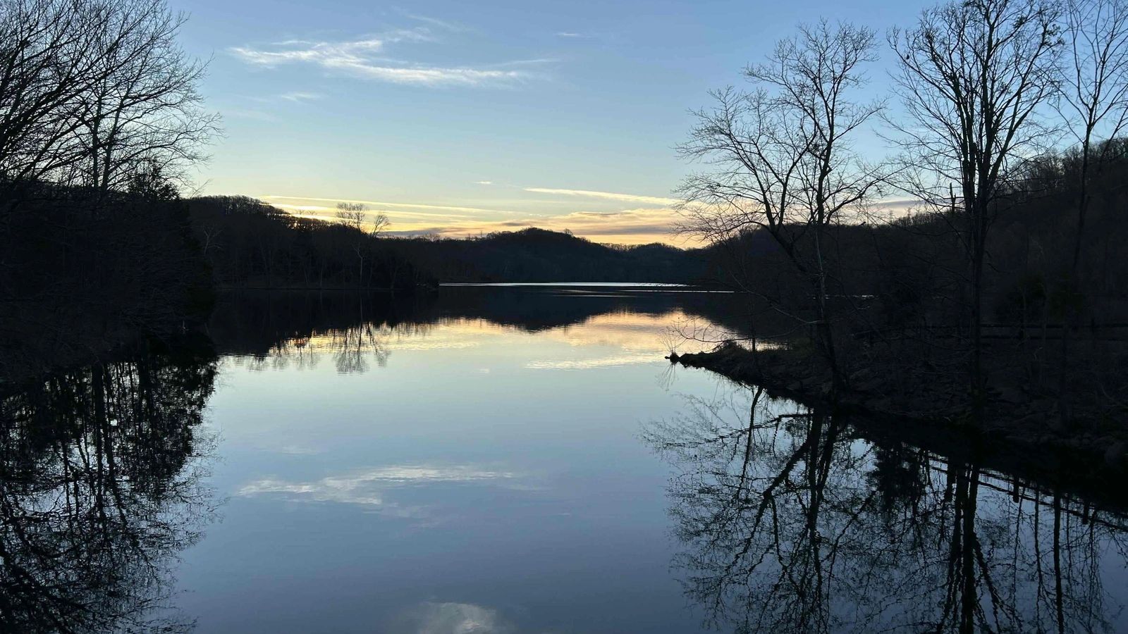 Calm lake at dusk reflecting bare trees and a pale evening sky