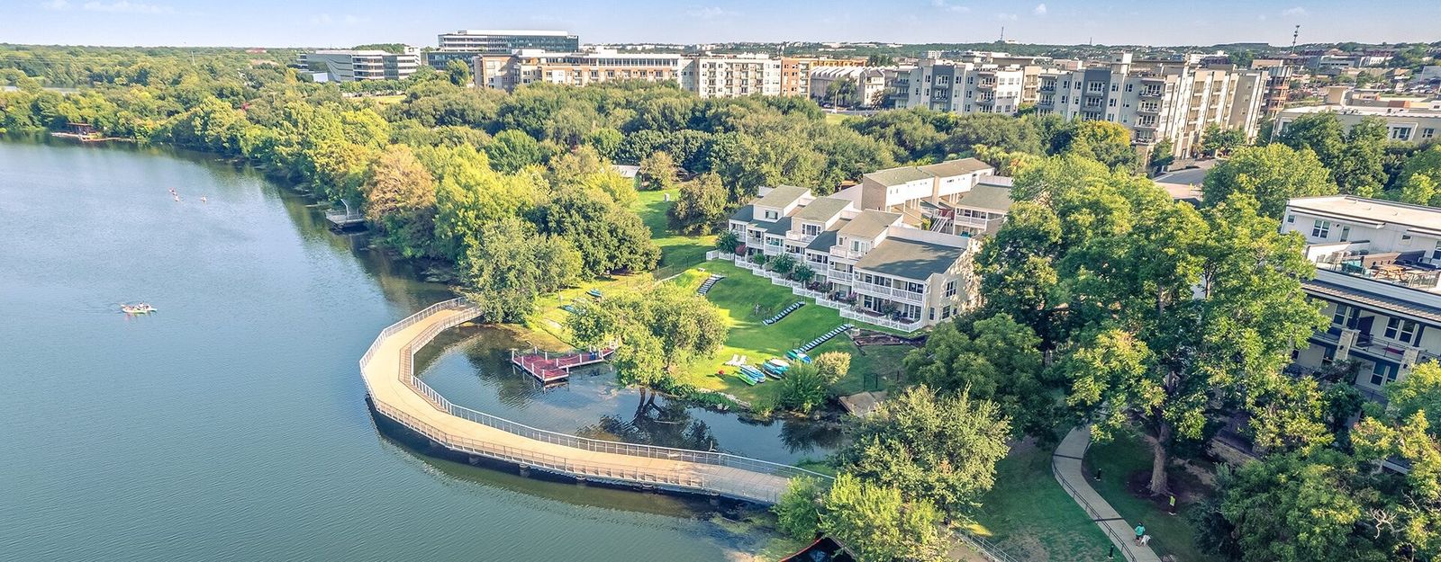 An aerial view of a curved boardwalk over a lake beside apartment buildings and trees.