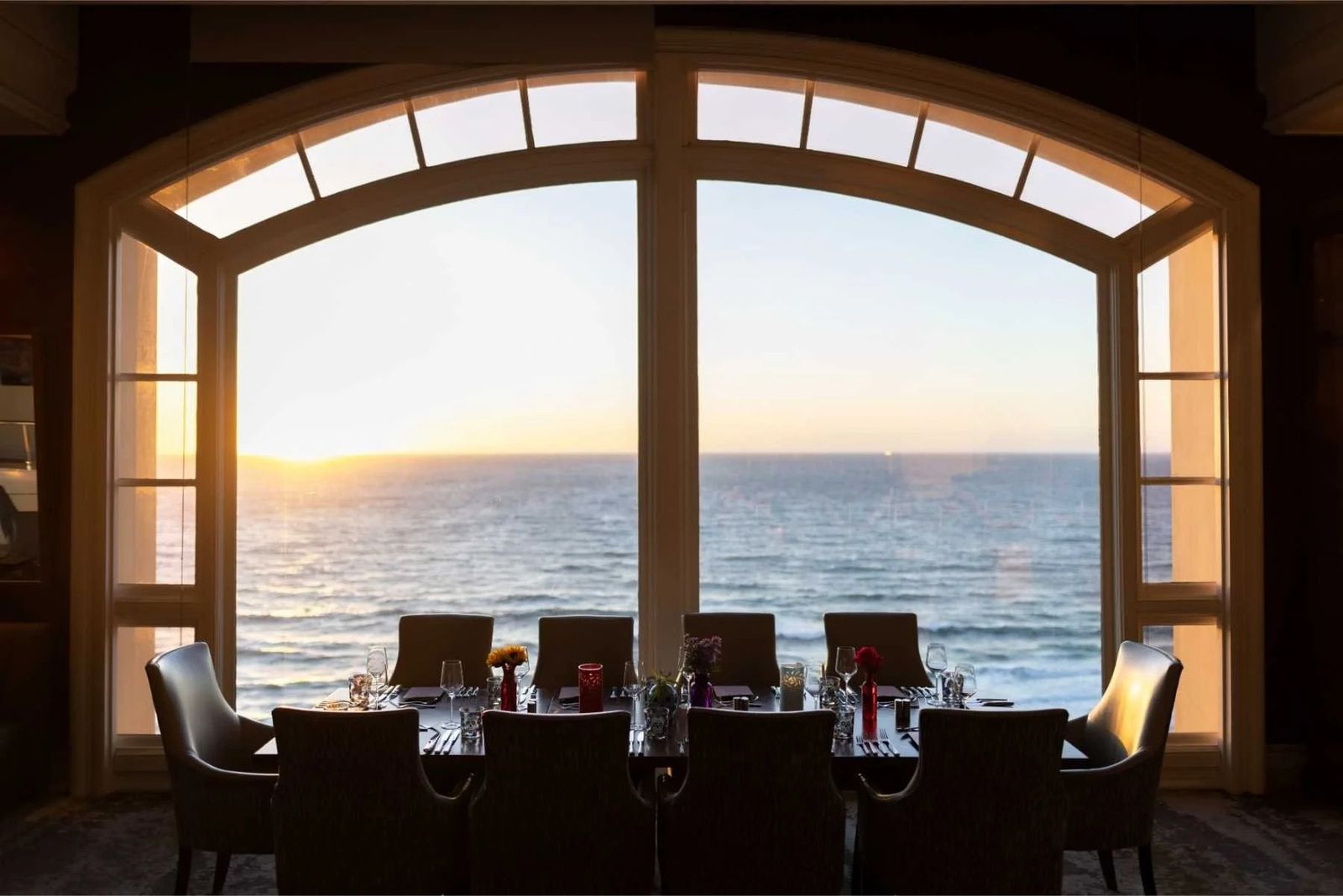 Dining tables in front of arched windows looking out at the ocean at sunset.