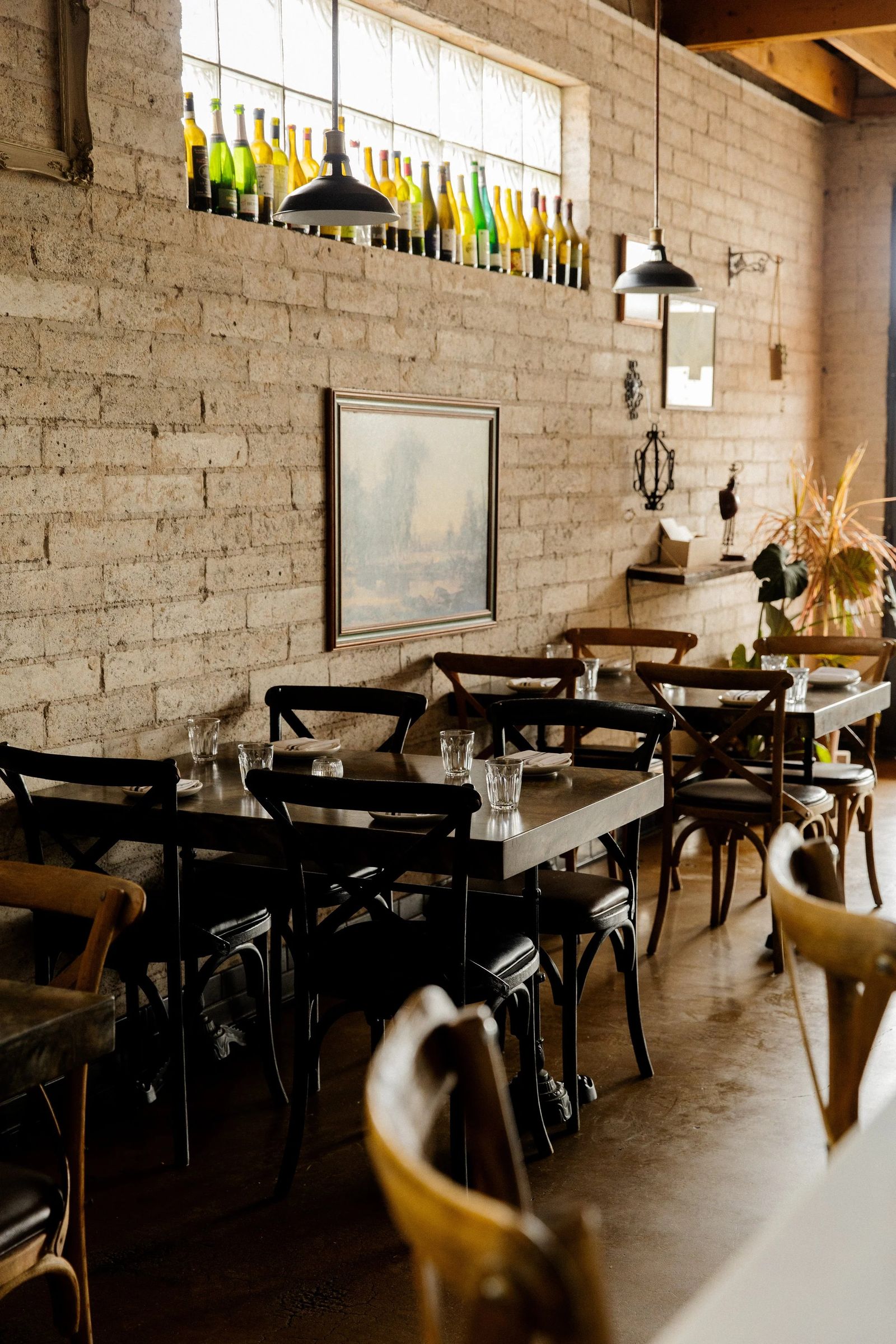 Dining room with a brick wall, black wooden chairs, small tables and a row of green bottles in the high windows