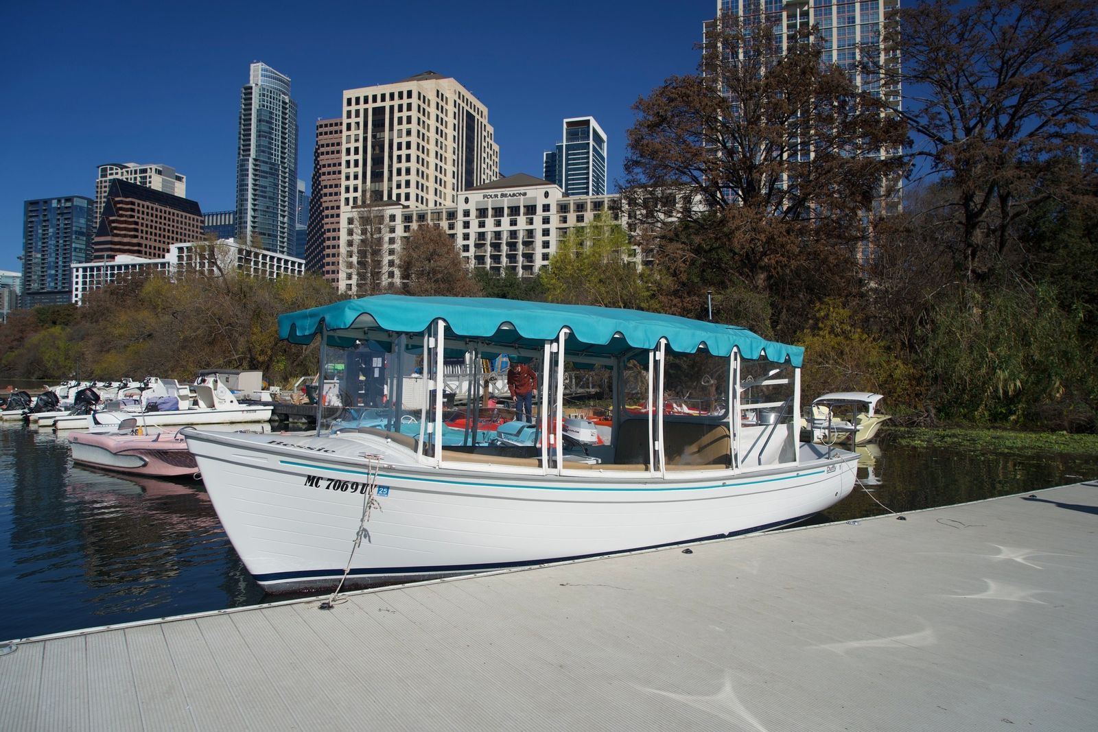A cream-and-teal canopy boat at a dock on a lake with city towers behind it.