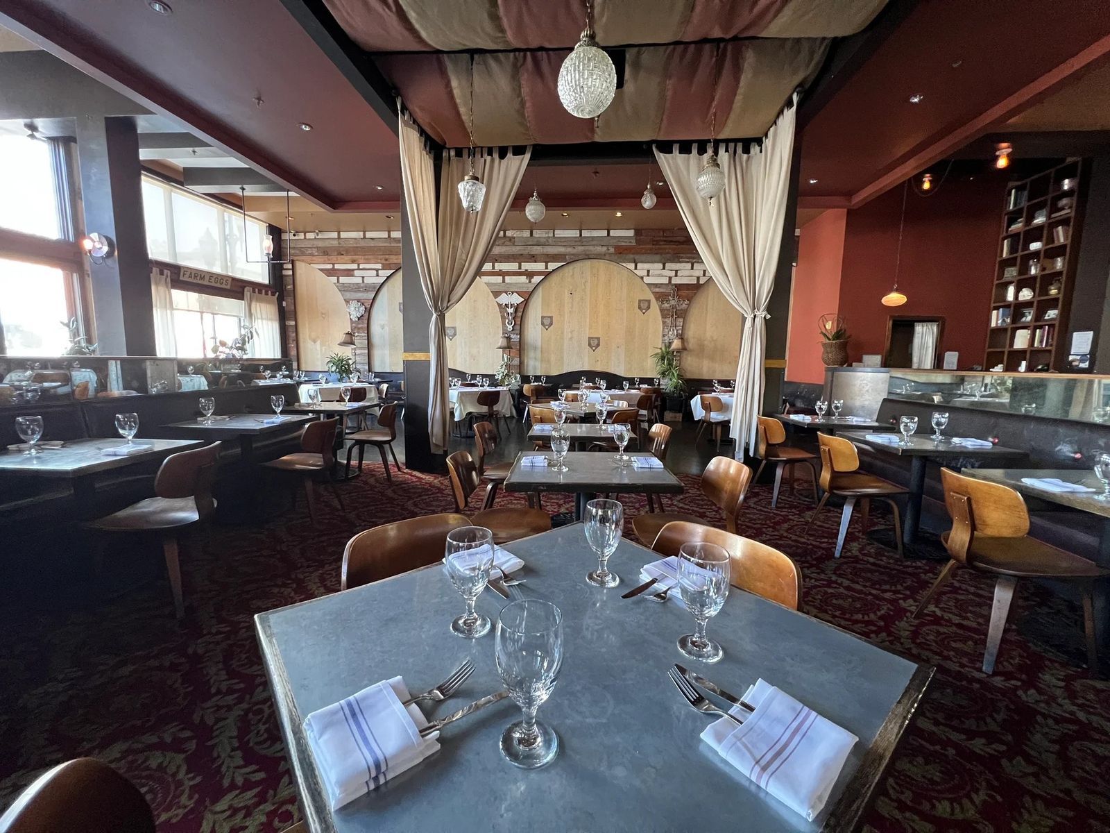 A dining room with zinc-topped tables, draped curtains and tall windows, with a bar beyond.