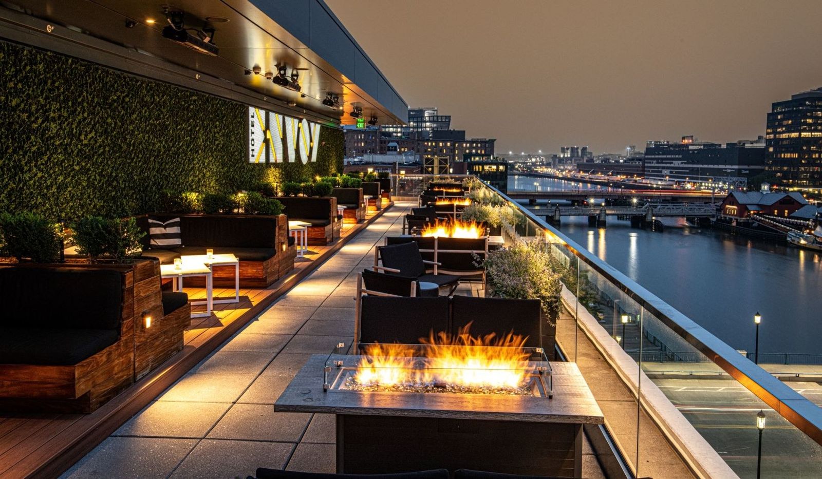 Rooftop terrace at dusk with fire pits, lounge seating and a view of the harbor